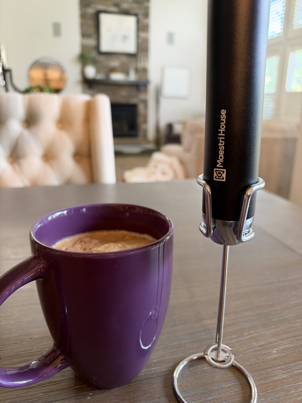 The Maestri House LunaFro Max in its chrome stand on a wooden table next to a purple mug of frothed coffee